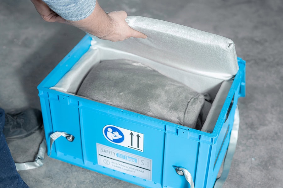 Uma mão abre uma caixa azul com uma almofada confortável, conteúdo cinzento, ambiente neutro.