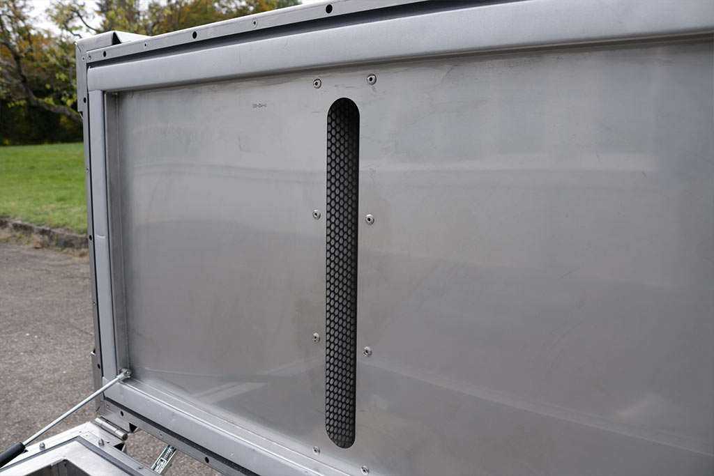 Painel metálico com grelha de ventilação preta de malha fina, rodeada por uma moldura prateada.