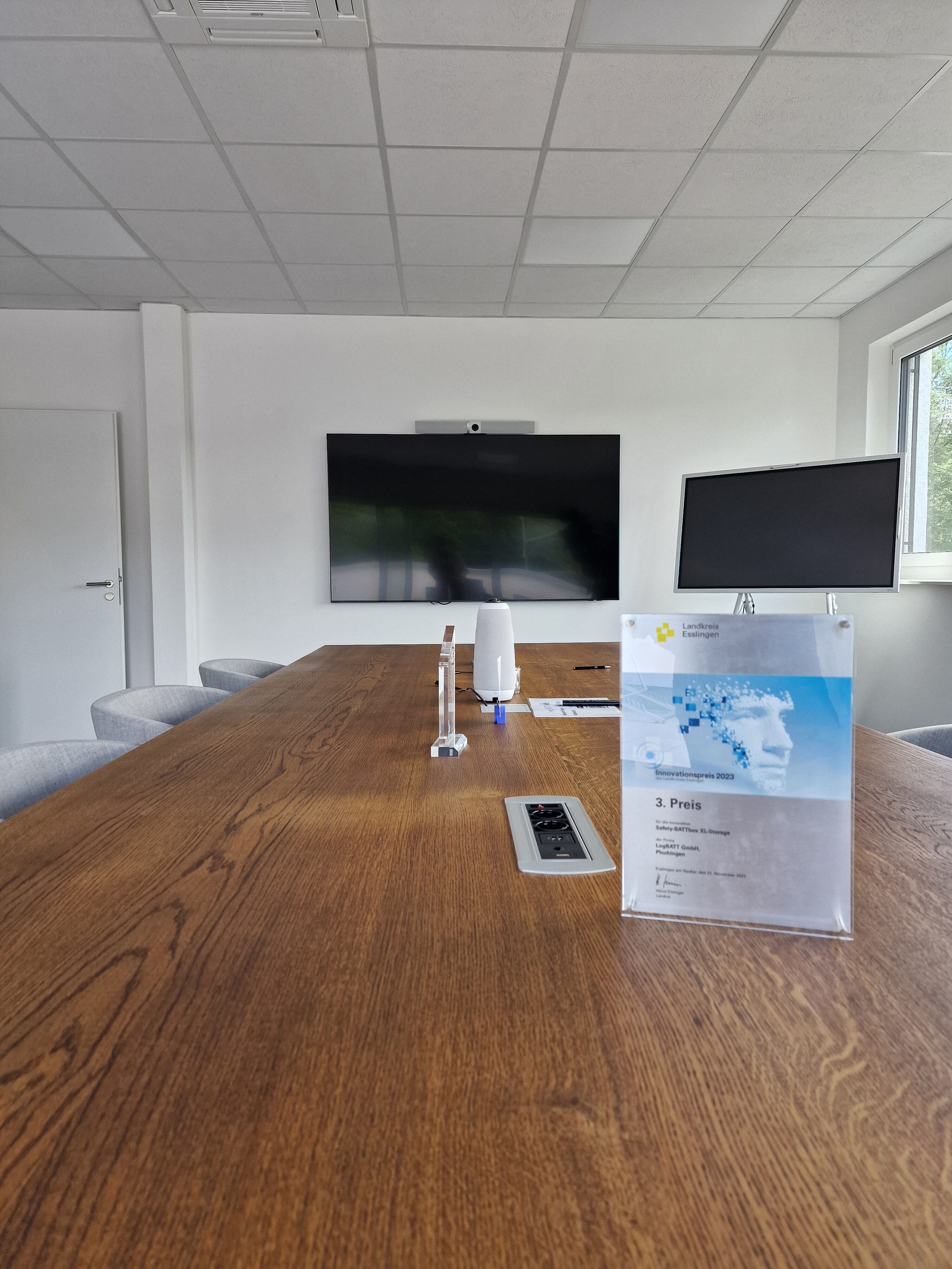 Sala de reuniões moderna com uma grande mesa de madeira, ecrã e cadeiras cinzentas. Ambiente luminoso.